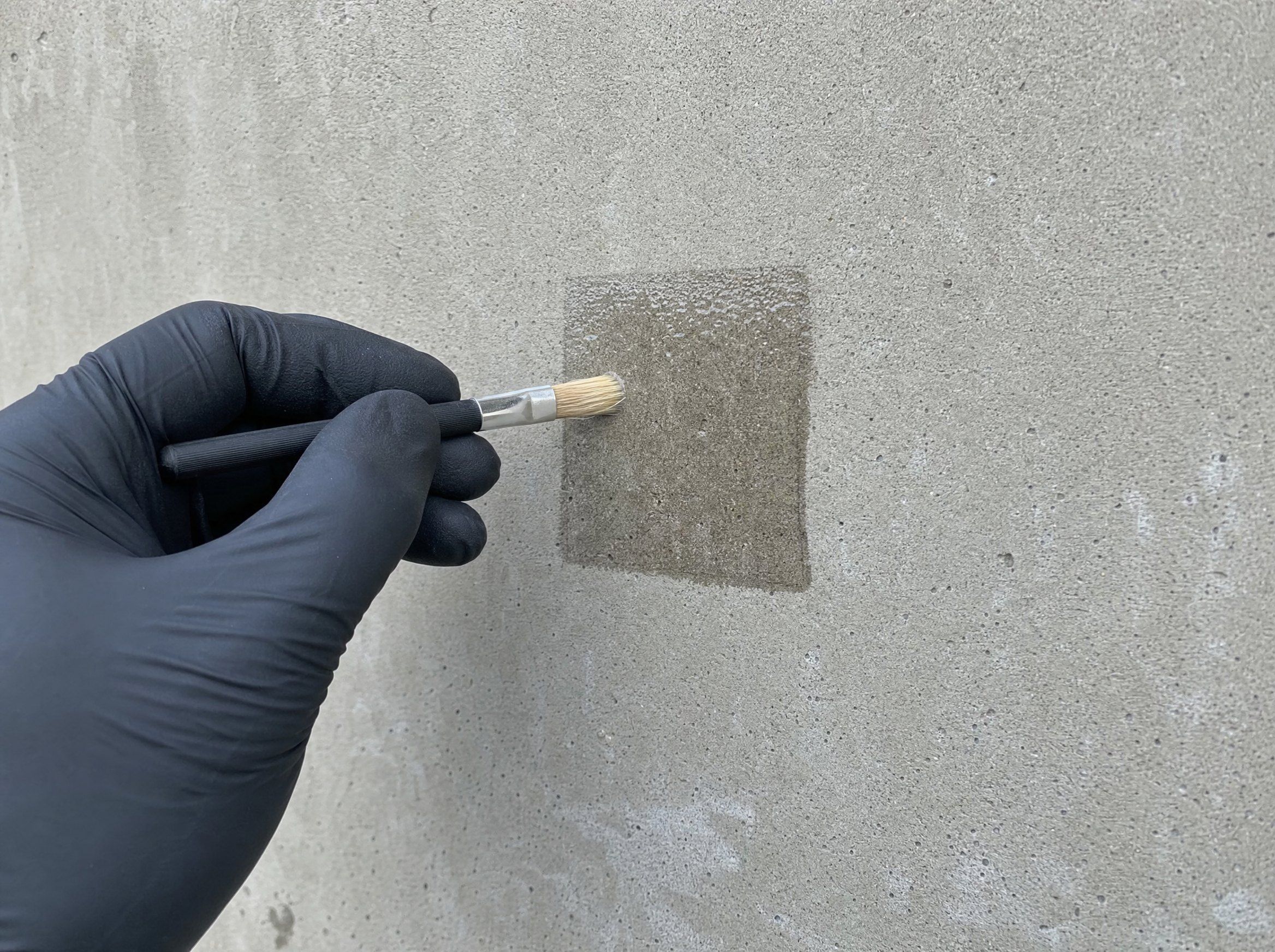 Behandschuhte Hand legt mit einem Pinsel eine kleine Probefläche auf Beton an